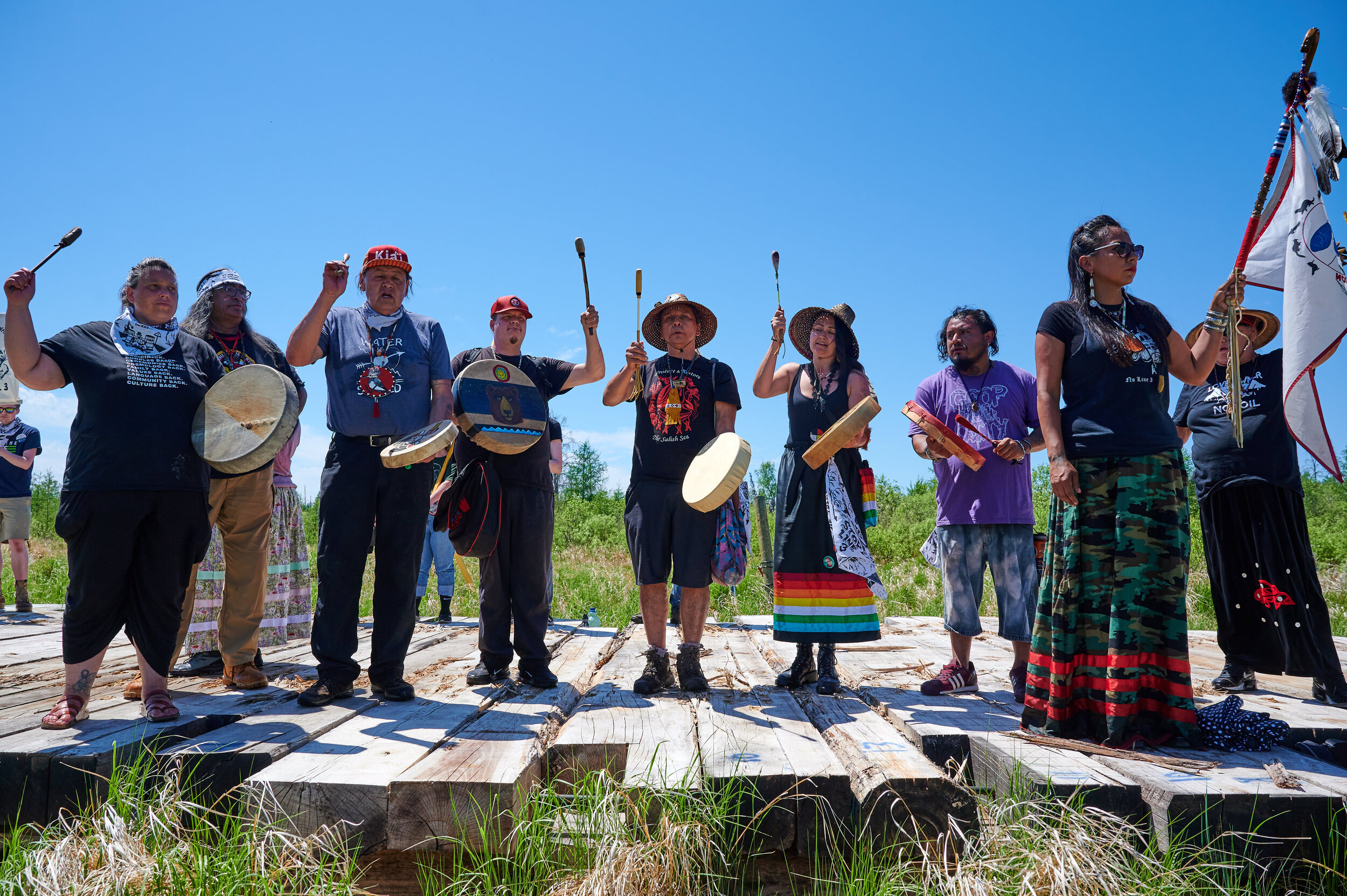 Water protectors and community members holding hand drums and singing on a wooden platform at a pipeline resistance action